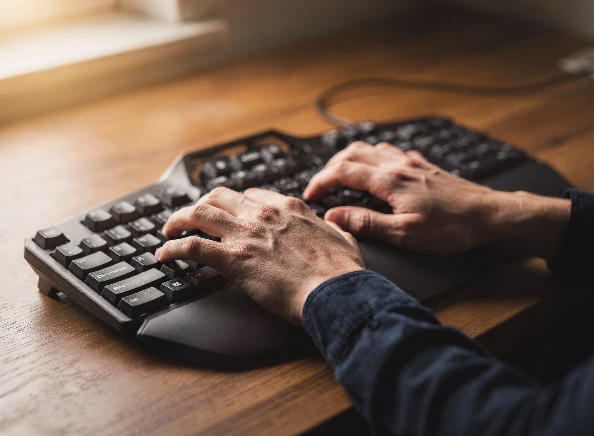 Hands typing on a black split ergonomic keyboard with integrated palm rests on a warm wooden desk.