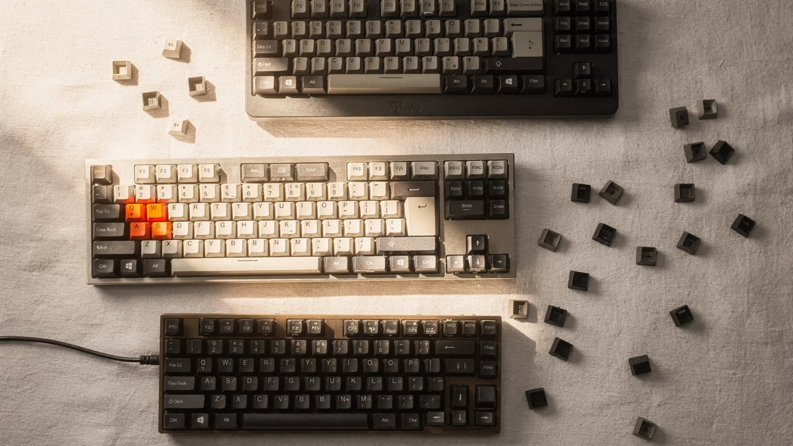 Three compact mechanical keyboards on a textured desk with loose keycaps around them